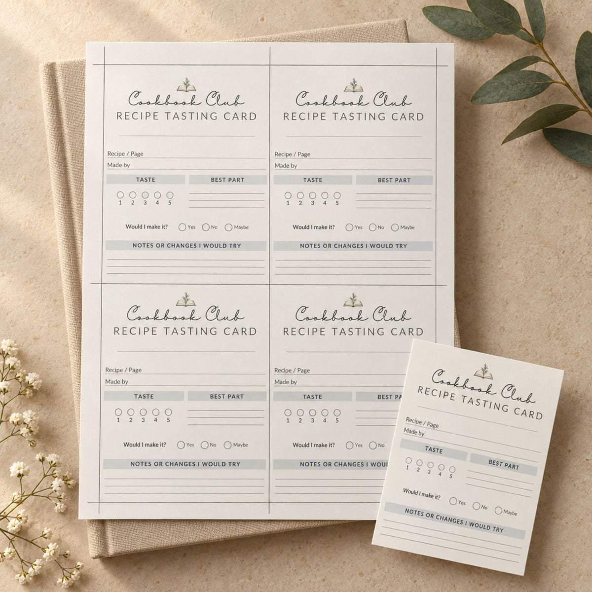 Four identical recipe tasting cards arranged on a beige surface with a folded copy in front, styled with dried flowers and green leaves. The cards are white with gray text and a small decorative illustration at the top.